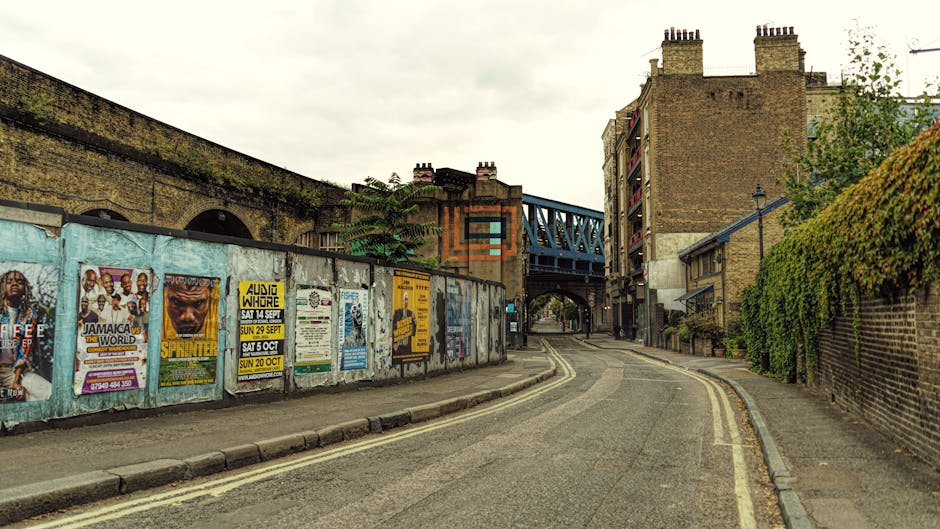 A narrow street in Walworth with a gentle right curve, showing an urban environment with a brick and concrete wall on the left, covered in colorful posters advertising events and concerts, and a brick wall with green foliage on the right. The road has double yellow lines along the curb, and a blue metal bridge spans above the street near the distance. The scene is lit with natural daylight under an overcast sky. This setting illustrates an area where home relocation or furniture transport might occur, with the street suitable for vehicle access and loading for local removals services, such as those offered by Man with Van Walworth. Visible elements include street pavement, storefront walls, and typical city architecture in the background.
