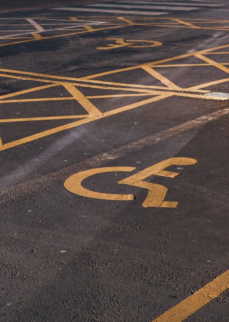 A close-up view of a parking lot surface features multiple yellow-painted parking bays, including marked disabled spaces with the international wheelchair symbol. The asphalt appears slightly worn with visible patches and tire marks. The yellow lines and symbols are clearly delineated, indicating designated parking areas for accessible parking. The image captures part of the parking lot, with the disabled parking section positioned at an angle, suggesting the area is near a building entrance or loading zone typically used for home relocations or furniture transport by Man with Van Walworth. No vehicles, furniture, or moving equipment are visible in the image; it solely highlights the accessibility markings on the ground, suitable for understanding parking arrangements for moving or relocation services.