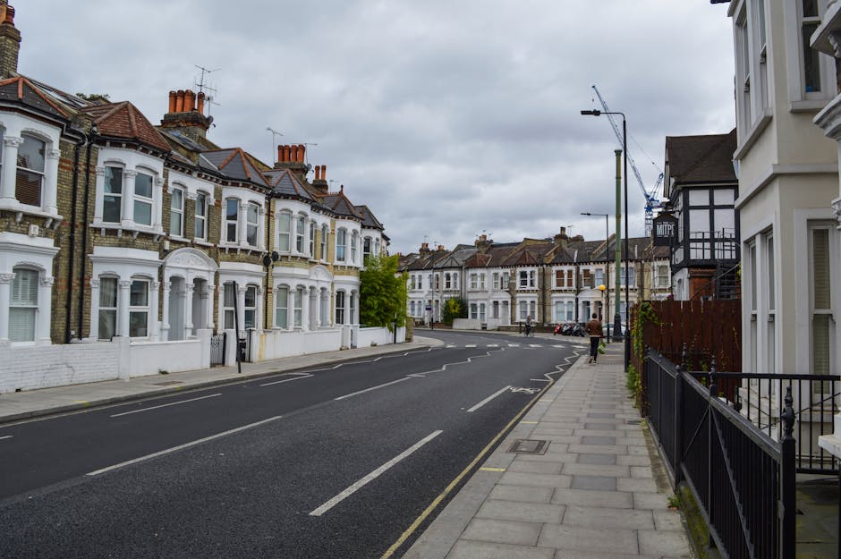 A narrow street in Walworth with a gentle right curve, showing an urban environment with a brick and concrete wall on the left, covered in colorful posters advertising events and concerts, and a brick wall with green foliage on the right. The road has double yellow lines along the curb, and a blue metal bridge spans above the street near the distance. The scene is lit with natural daylight under an overcast sky. This setting illustrates an area where home relocation or furniture transport might occur, with the street suitable for vehicle access and loading for local removals services, such as those offered by Man with Van Walworth. Visible elements include street pavement, storefront walls, and typical city architecture in the background.