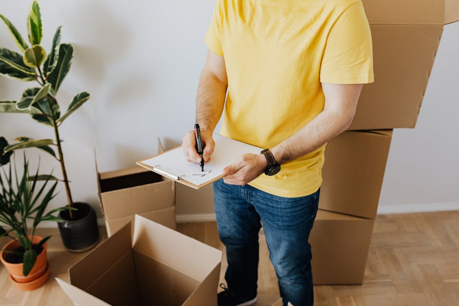 A person wearing a yellow t-shirt and blue jeans is inside a room, holding a clipboard and marker, appearing to record details during a home relocation process. The individual is standing near several open cardboard boxes and a stack of sealed boxes, which are likely packed for a move. To the left, there are potted houseplants on the floor, and the background features a white wall. The scene takes place in natural light, with the person engaged in planning or inventory checks related to furniture transport and packing during a house removal. This visual aligns with services offered by Man with Van Walworth, who specialise in furniture transport and relocation logistics, as shown on the Aylesbury Estate Removals page.