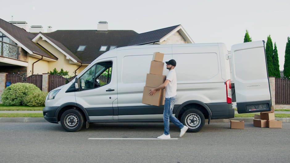 A bright blue vintage-style van parked on a sidewalk in front of a large beige brick wall with visible stains and cracks, beneath a dark grey horizontal panel. The van is situated behind a low metal f
