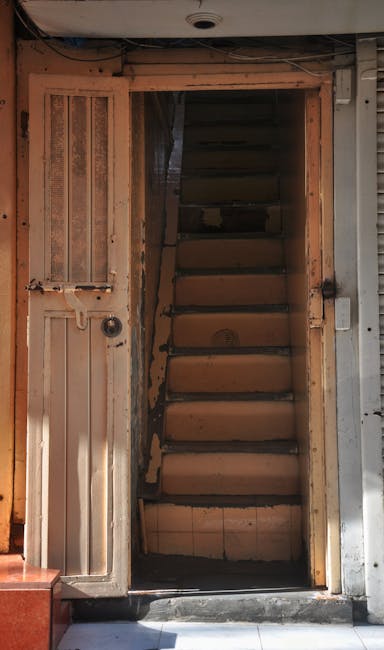 Inside a narrow staircase doorway during a home relocation, showing a compact wooden staircase with eight visible steps leading upwards, framed by a wooden doorframe, with the surrounding wall partially exposed and unfinished. The staircase is situated within a small, enclosed space, with some natural light illuminating the area. To the left of the doorframe, there is an old, weathered wooden panel with a small latch, and the floor at the base consists of tiled flooring with a slight shadow cast by the doorway's edge. Nearby, to the right, part of a metallic storefront shutter and wall panel are visible, indicating the proximity of a commercial area or building entrance. The environment suggests the initial stage of furniture transport and packing in a property with narrow Victorian stairs, as handled by Man with Van Walworth’s professional removals service, illustrating the challenges of navigating tight staircases during house moves in SE17.