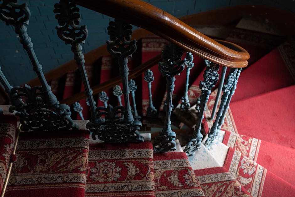 A view of a narrow Victorian staircase inside a property in Walworth, showing detailed black cast iron balustrades with ornate floral patterns, a polished wooden handrail on top, and a red patterned carpet runner covering the stairs. The staircase is situated underneath a low ceiling, and the area is illuminated by soft lighting. The image captures the space during a home relocation process, with the painted brick wall visible in the background. This setting reflects the challenges of furniture transport and packing through tight, historic stairs, with a focus on careful handling and careful planning by Man with Van Walworth, a professional removals service specializing in residential moves in SE17.