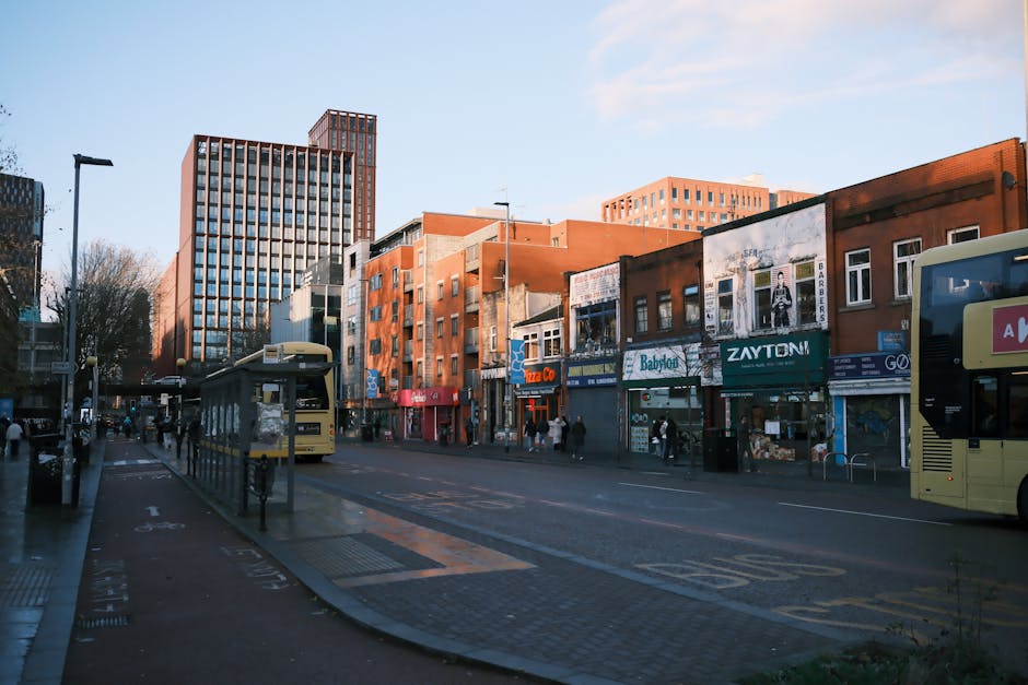 A city street scene during daytime with a modern high-rise building on the left and several low-rise commercial buildings on the right, featuring storefronts such as a pizzeria, a baby shop, and retail shops with signage. The street is populated with pedestrians walking on sidewalks and waiting at bus stops, with two double-decker buses present, one on each side of the road. The road includes designated lanes and cycling paths, with visible road markings and street lighting. The environment is clear, with some leafless trees indicating an autumn or winter season. This scene depicts urban transportation and shopping activity typical of a city centre, aligning with the services of Man with Van Walworth for house removals, furniture transport, and local moving logistics within the area near Elephant & Castle to Walworth.