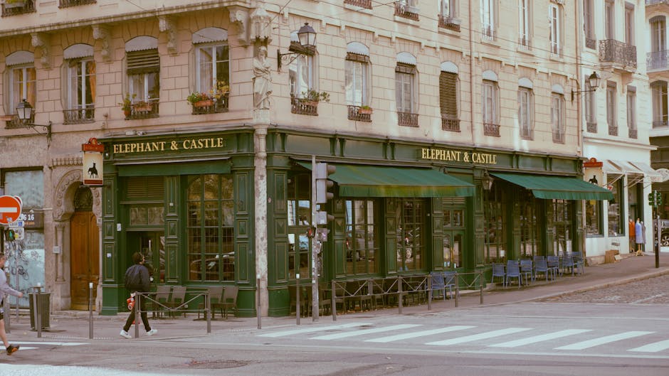 A street scene outside the Elephant & Castle pub in Walworth showing the pub's exterior with dark green painted wood paneling and large windows, some of which are covered with green fabric awnings. The building is part of a multi-story, light-colored stone London townhouse with decorative window frames and small balconies. In front of the pub, there are outdoor seating arrangements with several empty chairs and tables on the sidewalk, protected by low metal barriers. Pedestrians are walking along the pavement, including a person carrying a bag and another pushing a bicycle. The street features a pedestrian crossing with white zebra stripes, a street lamp, and traffic signs, including a no-entry sign on the corner. The scene captures a typical day with natural daylight, and it may be part of a moving or home relocation context, as a professional removals company like Man with Van Walworth could be involved in house removals or furniture transport from this location.