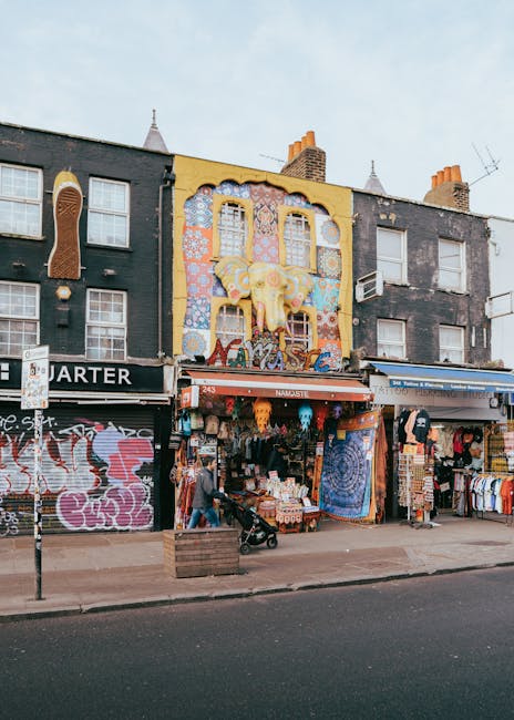 A city street scene during daytime with a modern high-rise building on the left and several low-rise commercial buildings on the right, featuring storefronts such as a pizzeria, a baby shop, and retail shops with signage. The street is populated with pedestrians walking on sidewalks and waiting at bus stops, with two double-decker buses present, one on each side of the road. The road includes designated lanes and cycling paths, with visible road markings and street lighting. The environment is clear, with some leafless trees indicating an autumn or winter season. This scene depicts urban transportation and shopping activity typical of a city centre, aligning with the services of Man with Van Walworth for house removals, furniture transport, and local moving logistics within the area near Elephant & Castle to Walworth.
