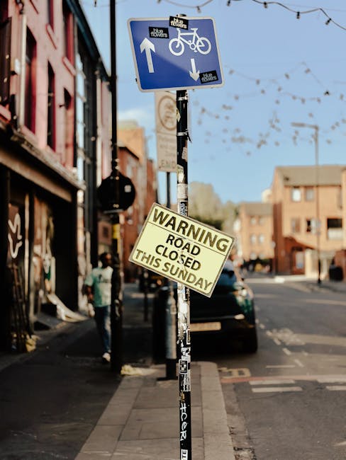 A street scene featuring a black metal pole with two signs attached. The upper sign is blue with white symbols indicating a shared pathway for pedestrians and cyclists, with arrows pointing in opposite directions. Below, a yellow warning sign states, 'ROAD CLOSED THIS SUNDAY.' The pole is on the sidewalk, with buildings and shops visible on the left side and parked vehicles along the curb. In the background, a black van is parked adjacent to the pole, and there are pedestrians walking along the pavement. The scene suggests a period when road closures are scheduled, which could impact house removals or furniture transport logistics, especially for businesses like Man with Van Walworth involved in home relocation and packing and moving services in the area.