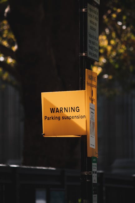 A yellow rectangular warning sign attached to a metal pole displays the message 'WARNING Parking suspension' in black text, with additional smaller text below that is not fully visible. The sign is situated outdoors, with a blurred background featuring a large tree, a building, and a fence, indicating an urban setting. The sign is illuminated slightly by natural light, suggesting daytime. This signage relates to road closures and parking suspensions that may impact house removals or furniture transport services, which are often coordinated by companies like Man with Van Walworth during relocation projects within the Walworth area. The pole appears to have additional notices or parking information attached above and below the warning sign, emphasizing the importance of checking for parking restrictions before moving day, to ensure a smooth loading process when relocating furniture or household items.