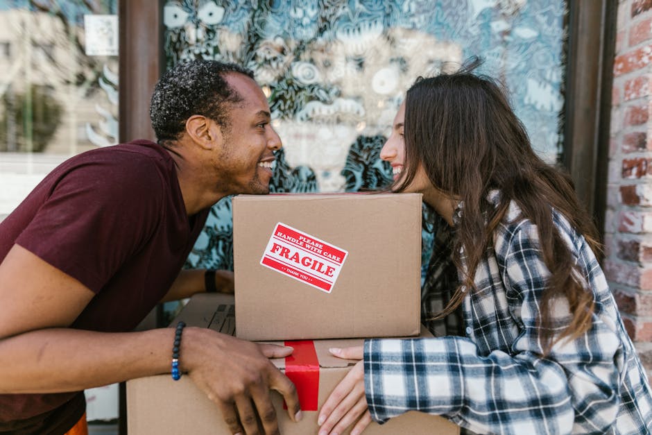 A man and woman are engaged in a home relocation activity outdoors near a building with a glass window and brick wall. The man, of African descent with curly hair, wears a maroon T-shirt and is smiling while lifting a cardboard box labeled 'Fragile' with a red and white handling sticker. The woman, with long brown hair and wearing a plaid shirt, faces him and is also smiling as she supports the boxes. The boxes, wrapped in plastic and placed inside a moving area, appear to be part of a packing and moving process. In the background, there are some furniture blankets and a moving van with its door open, ready for loading. The scene captures the careful handling of household items during a furniture transport or home packing operation, illustrating the operational aspects of a professional removals service like Man with Van Walworth.