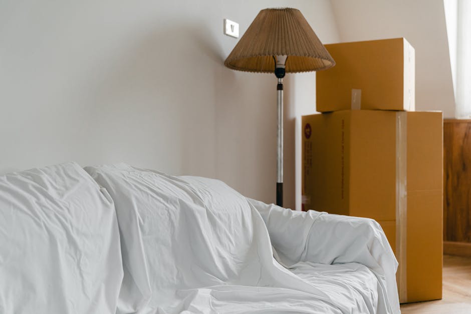 Inside a room with cream-colored walls and wooden flooring, there are three large cardboard boxes stacked against the wall, with the smallest box placed on top of the other two. To the left, a white fabric-covered sofa with visible creases and folds is positioned close to the wall. A tall standing floor lamp with a brown pleated lampshade and a metallic silver stand is situated near the boxes, casting soft shadows on the wall. The scene appears to be part of a home relocation or packing process, with the boxes prepared for moving. The room is lit by natural light coming through a window on the right side, partially visible, creating a bright and neutral environment. The image indicates a moving company's involvement, such as Man with Van Walworth, handling packing and furniture transport during a house removal, emphasizing the packing and loading stages of the relocation process.
