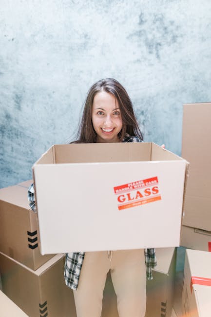 A man and woman are engaged in a home relocation activity outdoors near a building with a glass window and brick wall. The man, of African descent with curly hair, wears a maroon T-shirt and is smiling while lifting a cardboard box labeled 'Fragile' with a red and white handling sticker. The woman, with long brown hair and wearing a plaid shirt, faces him and is also smiling as she supports the boxes. The boxes, wrapped in plastic and placed inside a moving area, appear to be part of a packing and moving process. In the background, there are some furniture blankets and a moving van with its door open, ready for loading. The scene captures the careful handling of household items during a furniture transport or home packing operation, illustrating the operational aspects of a professional removals service like Man with Van Walworth.
