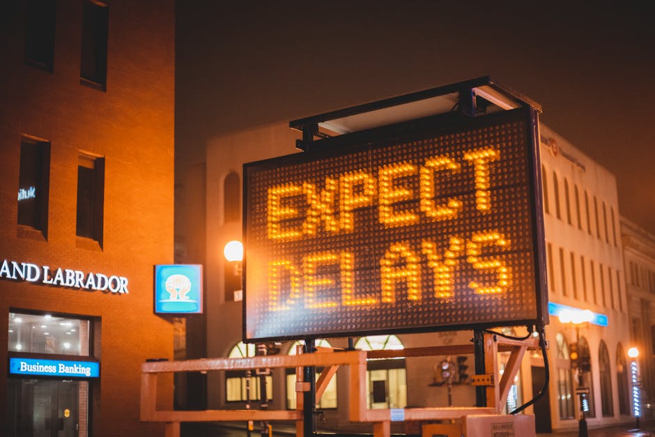 A large, illuminated electronic roadside sign displaying the message 'EXPECT DELAYS' in bright orange letters is mounted on a metal frame. The sign is situated on a city street at night, with surround