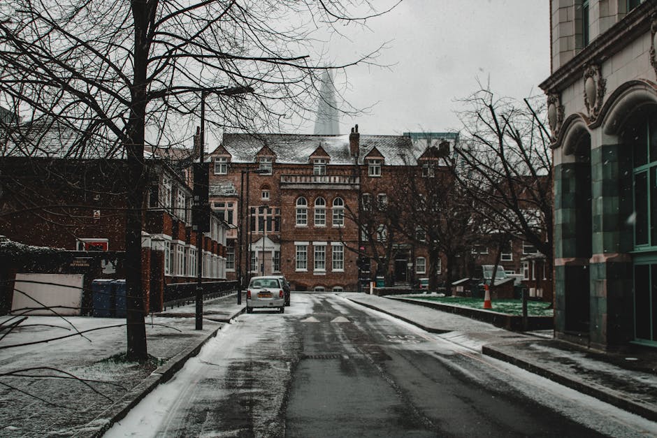 The image depicts a residential street during winter with a thin layer of snow covering the pavement and road, bordered by leafless trees and historic brick buildings, including a prominent multi-storey structure with arched windows and decorative brickwork. On the right side of the image, a narrow sidewalk runs alongside a modern storefront with large, curved glass windows. A small white van is parked on the street, near the curb, close to a black bollard, suggesting recent or ongoing loading or unloading activities associated with house or furniture removals. The street scene is illuminated by natural daylight under an overcast sky, typical of a cold, winter day in Southwark. This environment aligns with the logistical considerations of home relocation or furniture transport services, such as those offered by [COMPANY_NAME], operating within the context of Southwark Council’s permit requirements for removals on Walworth Road. The overall scene reflects the practical aspects of packing, loading, and delivering household items in an urban residential area during winter conditions, supporting efficient moving and transport operations.