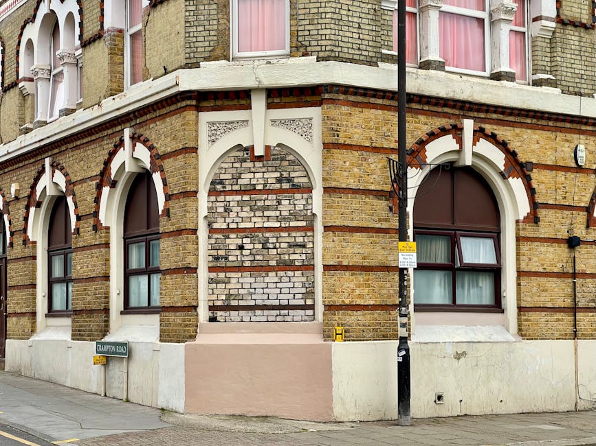 Photo of a corner of a Victorian-style brick building on Walworth Road, with arched windows featuring brown frames and pink curtains inside. The building has decorative white wooden trim and contrasting red brick accents around the arches. A black lamppost with a yellow and white parking restriction sign and a small yellow 'H' sign is located at the sidewalk corner. The pavement in front shows signs of recent work, with a section of pale pink painted wall near a small level entrance step. A street name sign indicates 'Crawford Road' on the corner. This setting may relate to house removals or property relocation activities, with the exterior environment and architectural details capturing the context of an urban house or flats in Southwark, relevant for professional furniture transport and moving services provided by Man with Van Walworth. The scene is captured in natural daylight, emphasizing the building's historic brickwork and structural details.