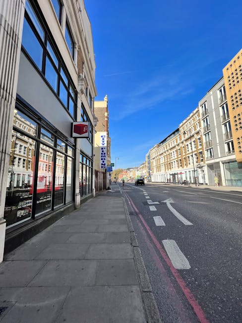 The image depicts a residential street during winter with a thin layer of snow covering the pavement and road, bordered by leafless trees and historic brick buildings, including a prominent multi-storey structure with arched windows and decorative brickwork. On the right side of the image, a narrow sidewalk runs alongside a modern storefront with large, curved glass windows. A small white van is parked on the street, near the curb, close to a black bollard, suggesting recent or ongoing loading or unloading activities associated with house or furniture removals. The street scene is illuminated by natural daylight under an overcast sky, typical of a cold, winter day in Southwark. This environment aligns with the logistical considerations of home relocation or furniture transport services, such as those offered by [COMPANY_NAME], operating within the context of Southwark Council’s permit requirements for removals on Walworth Road. The overall scene reflects the practical aspects of packing, loading, and delivering household items in an urban residential area during winter conditions, supporting efficient moving and transport operations.