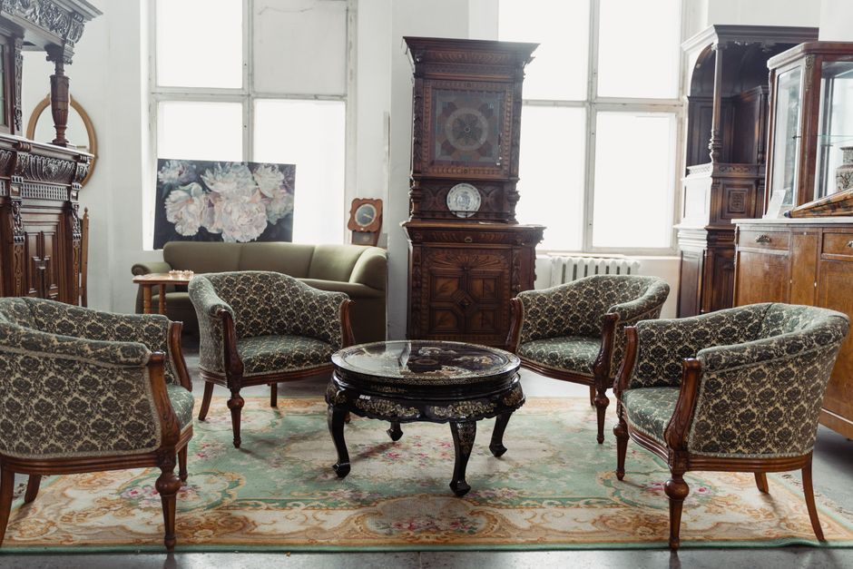 A well-lit room with large windows allowing natural light, containing antique wooden furniture including an upright clock, display cabinets, and a sideboard. In the foreground, four upholstered armchairs with patterned fabric and wooden frames are arranged around a decorative, dark-finished wooden table with curved legs and a glossy surface. The furniture is positioned on a patterned area rug with floral motifs. Outside the windows, a radiator is visible beneath the window sills. The scene depicts a typical setting for moving or packing antique furniture as seen in professional house removals services, with the furniture carefully arranged, possibly in the process of being loaded for transport to another location. The setting is clean, organized, and shows the arrangement of household items prior to a furniture transport or home relocation operation, likely facilitated by a professional removal company such as Man with Van Walworth.