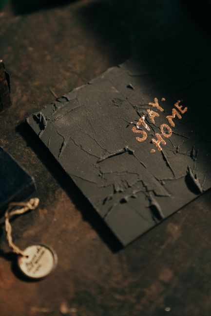 A black, textured cardboard box with a cracked and peeling surface, situated on a wooden floor inside a property during a house relocation. The box has the words 'SAME HOME' written in orange or brownish ink or marker on its top surface. To the left of the box, there is a small, round tag attached with a string, possibly indicating contents or used for identification. The background includes part of a dark-colored floor and some indistinct objects, suggesting a moving or packing process. The scene is lit with soft, natural light, and the box appears to be part of the packing materials used by [COMPANY_NAME], Man with Van Walworth, during furniture transport or home relocation activities, emphasizing careful handling and organized packing in the moving process.