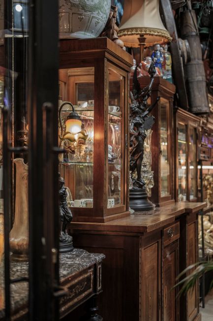 A well-lit room with large windows allowing natural light, containing antique wooden furniture including an upright clock, display cabinets, and a sideboard. In the foreground, four upholstered armchairs with patterned fabric and wooden frames are arranged around a decorative, dark-finished wooden table with curved legs and a glossy surface. The furniture is positioned on a patterned area rug with floral motifs. Outside the windows, a radiator is visible beneath the window sills. The scene depicts a typical setting for moving or packing antique furniture as seen in professional house removals services, with the furniture carefully arranged, possibly in the process of being loaded for transport to another location. The setting is clean, organized, and shows the arrangement of household items prior to a furniture transport or home relocation operation, likely facilitated by a professional removal company such as Man with Van Walworth.