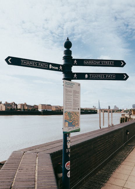 A black metal fold-up street signpost situated on a paved walkway along the river Thames in Walworth. The signpost features two horizontal directional signs pointing to 'Thames Path' and 'Narrow Street', with symbols indicating pedestrian routes. Below the street signs, there is an informational map and additional notices displayed on a white rectangular plaque. In the background, the river Thames stretches across the image, with a cityscape of modern buildings, including the London skyline with the Shard visible on the horizon. To the right of the signpost, a bench runs parallel to the river, and the sky above is partly cloudy. The scene captures a typical outdoor setting that could be part of a house removal or moving process, as movement and logistics are implied by the signage indicating routes along narrow streets and the Thames Path, relevant to transport and relocation planning, as covered on [PAGE_TITLE]. Natural lighting highlights the scene's urban environment and outdoor context, supporting the focus on outdoor logistics involved in furniture transport or house relocations. Man with Van Walworth occasionally supports content related to removals and moving services in this area.