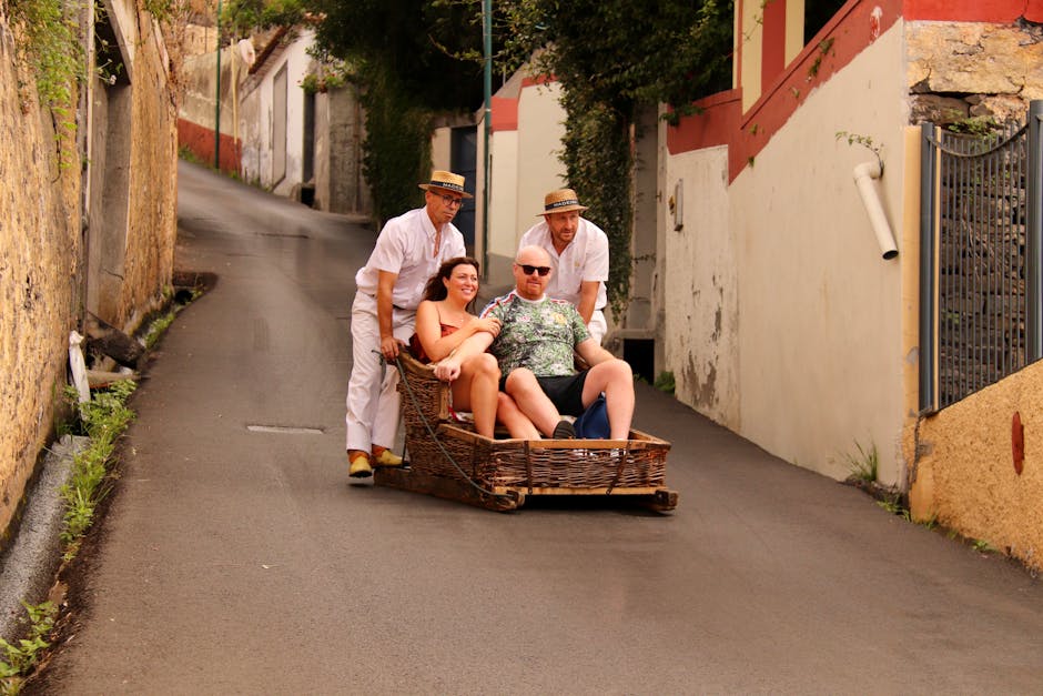 A humorous scene of a woman and a man sitting in a rectangular wooden basket on a narrow, sloped street. Two men dressed in white, wearing straw boater hats, are pushing the basket from behind; one is positioned at the rear, and the other at the front, both guiding the load. The woman has long dark hair and is smiling, while the man is bald, wearing sunglasses, and appears relaxed. The street is paved with dark asphalt, bordered by high, uneven stone and plaster walls painted in shades of white, cream, and red. Green plants grow along the edges of the walls and near the road surface. The setting appears to be in a quaint, older neighbourhood with closely packed houses and narrow passageways, creating a charming backdrop suggestive of house relocation or unusual furniture transport seen in relocation services by Man with Van Walworth. The scene is well-lit by natural daylight, emphasizing the casual yet practical atmosphere of furniture or household item moving in a residential area.